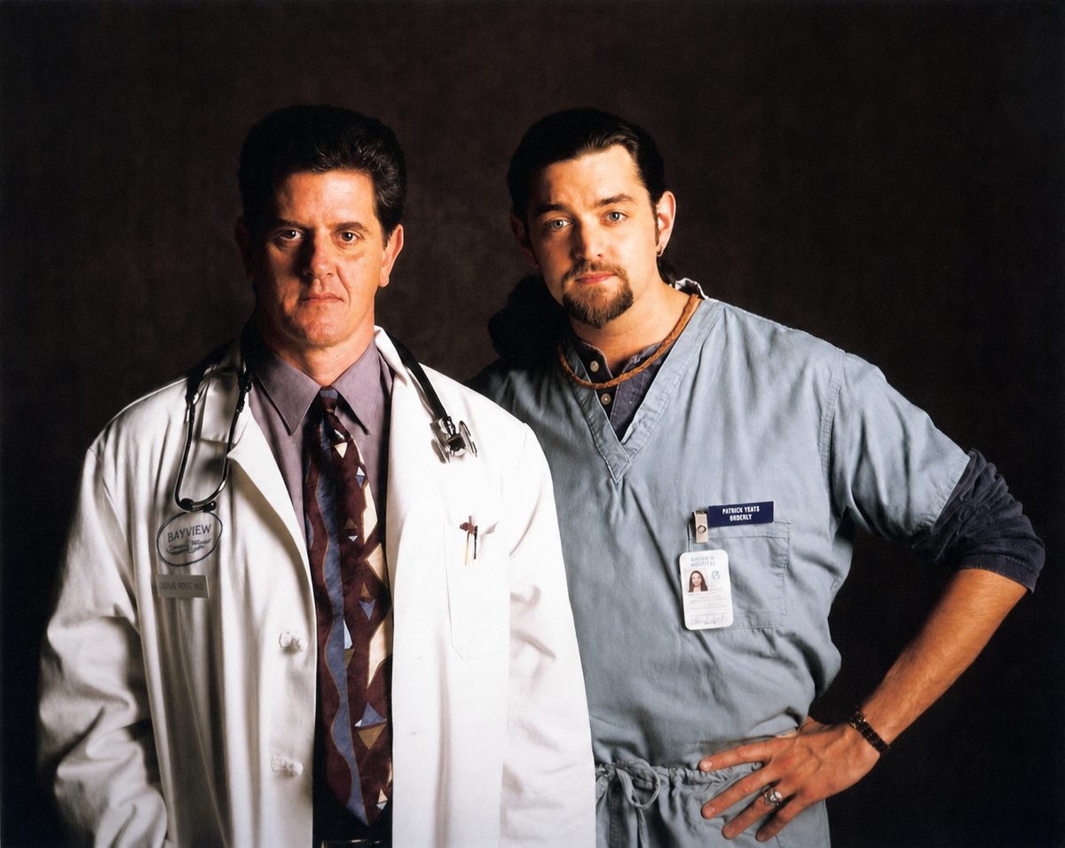 Sam McMurray in a doctor's coat and stethoscope beside a hospital orderly.